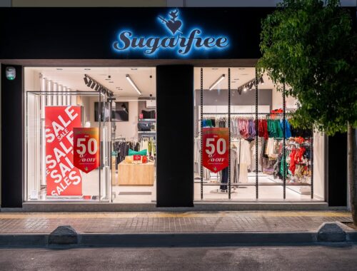 Ecocreta Construction Sugarfree - Storefront with neon sign, large sale banners advertising 50% off, and colorful clothing displayed on racks inside retail shop window.