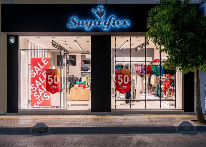 Ecocreta Construction Sugarfree - Storefront with neon sign, large sale banners advertising 50% off, and colorful clothing displayed on racks inside retail shop window.